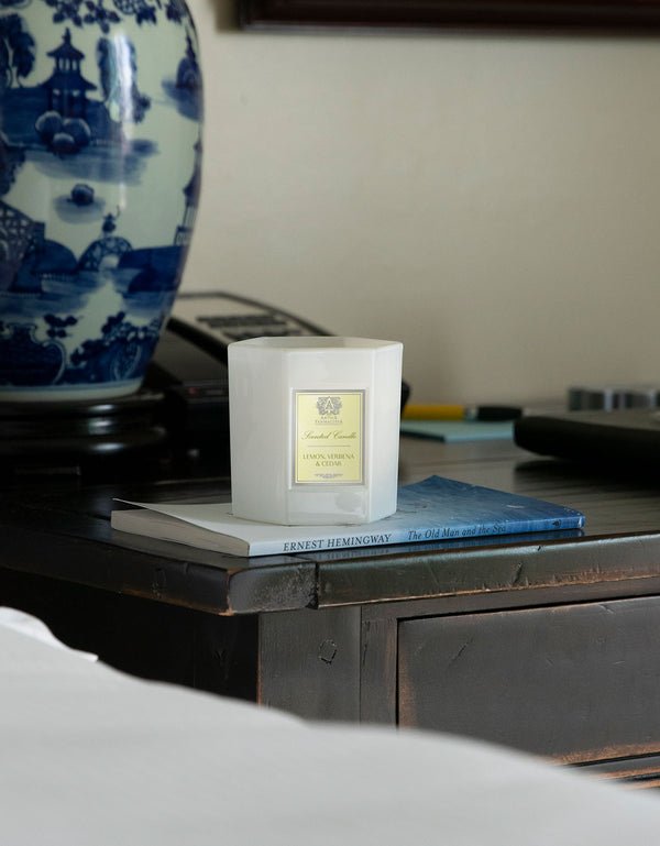 The Antica Farmacista Lemon Verbena & Cedar Candle rests on a closed copy of The Old Man and the Sea by Ernest Hemingway. The book is on a wooden nightstand beside a blue and white ceramic vase with traditional design, reminiscent of vintage apothecary bottles.