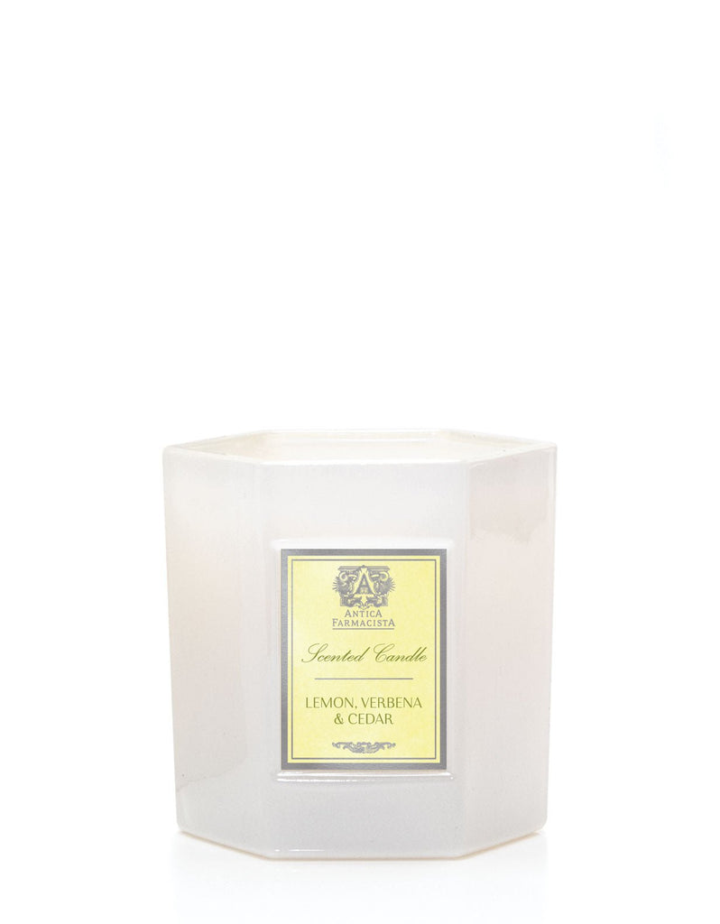 The Antica Farmacista - Lemon Verbena & Cedar Candle features a hexagonal frosted glass holder with an apothecary bottle charm. Its light yellow shade is enhanced by a decorative emblem in a darker hue above the text.