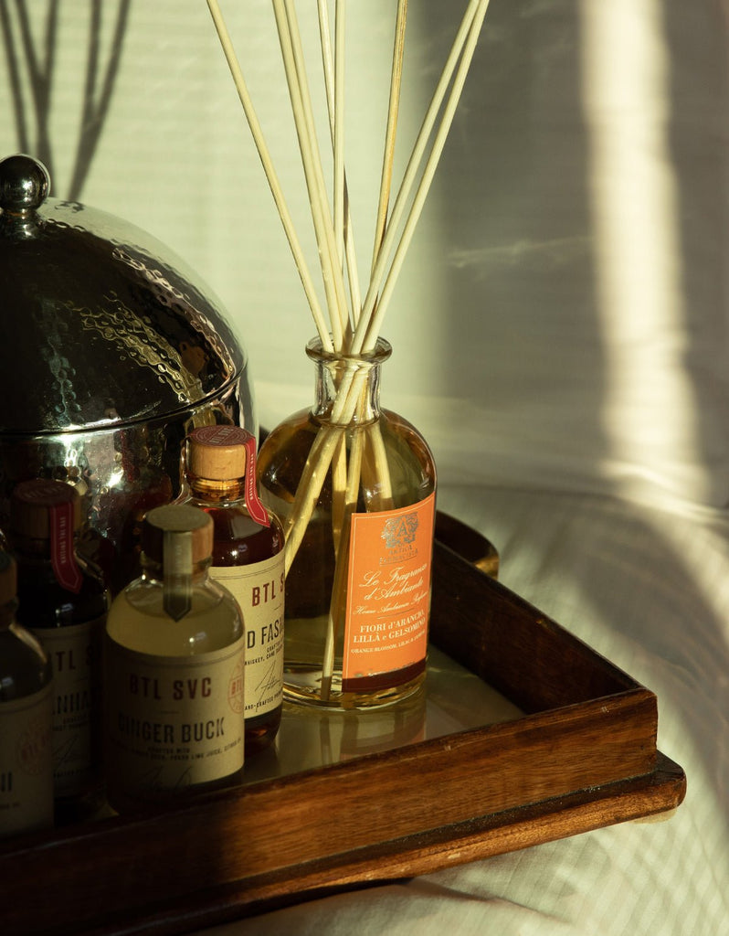 A wooden tray holds various bottles, including an Antica Farmacista reed diffuser labeled Orange Blossom, Lilac & Jasmine, with long reeds scenting the air. A metal ice bucket with a lid shares the space as sunlight casts gentle shadows.