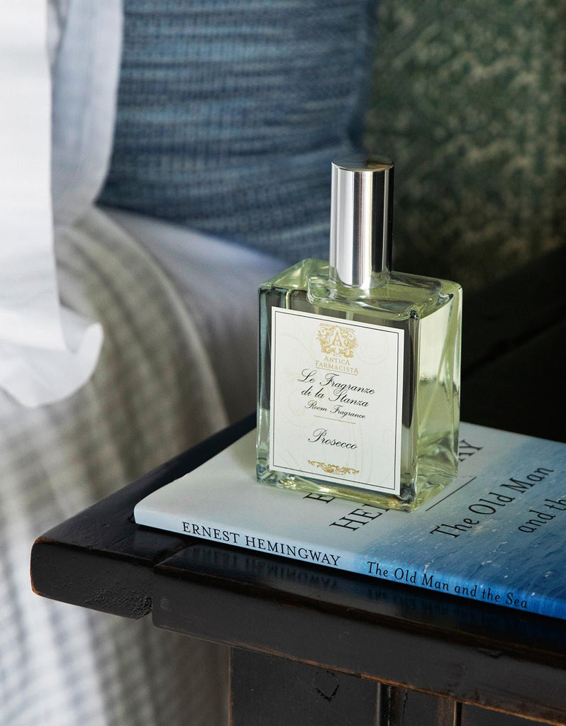 A Prosecco 100ml Room Spray by Antica Farmacista sits on a wooden surface near Hemingway’s “The Old Man and the Sea,” emitting a scent. The background includes a textured cushion and striped blanket.