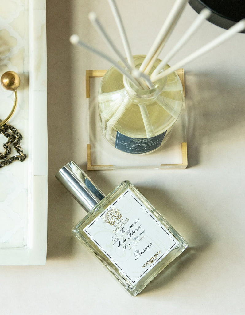 An Antica Farmacista silver-capped perfume bottle sits beside a reed diffuser, releasing Madagascar Vanilla scents. To the left is a decorative tray with jewelry, adding elegance to the scene.