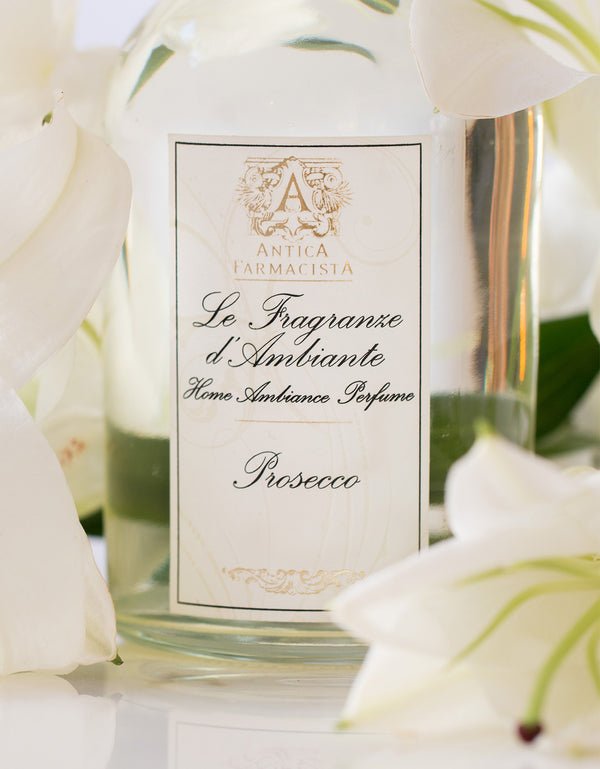 A 250ml Antica Farmacista Prosecco Reed Diffuser, surrounded by white lilies, features an elegant script label with a crest logo that captures its signature scent.