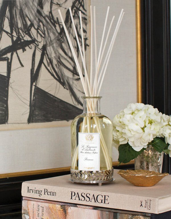 An Antica Farmacista Prosecco 250ml Reed Diffuser with white reeds rests atop books Irving Penn and Passage. Beside it, a small bowl and a vase of white hydrangeas complement the scene, while abstract black-and-white art adorns the background.