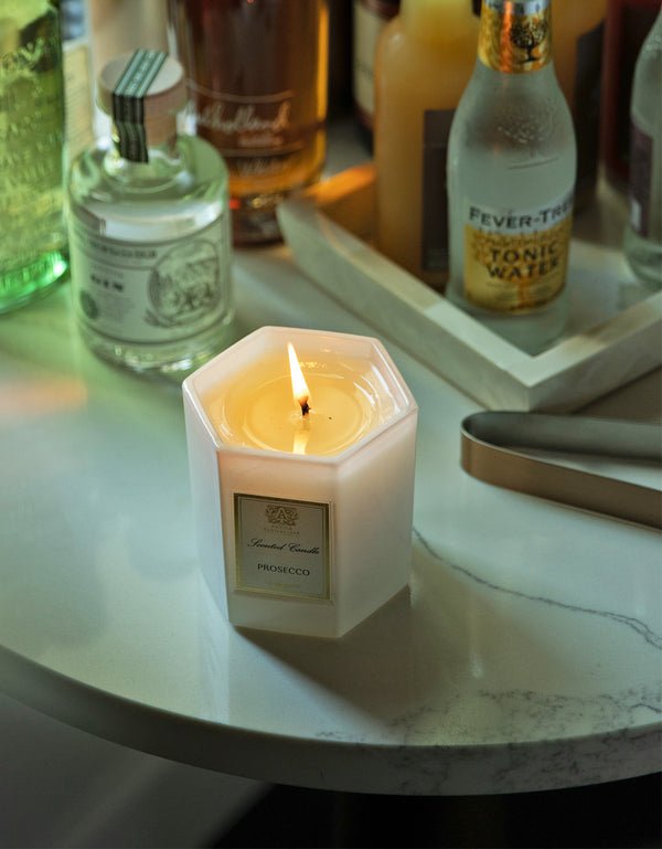 The Antica Farmacista - Prosecco 9oz Candle illuminates a marble surface, surrounded by glass bottles and a tray of tonic water. Its Satsuma Citrus aroma enhances the cozy and relaxed ambiance.