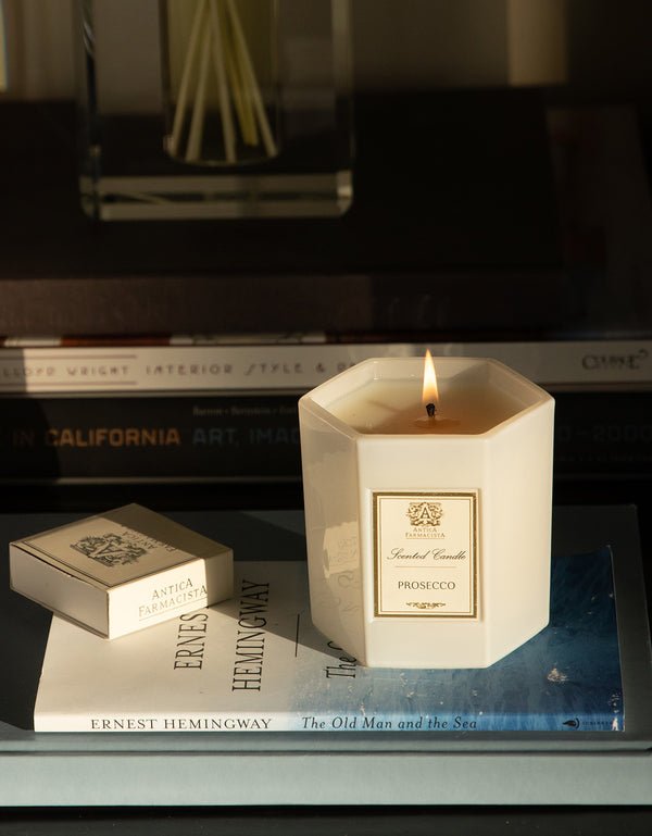 An Antica Farmacista - Prosecco 9oz Candle with Satsuma Citrus notes rests on The Old Man and the Sea by Ernest Hemingway, next to a box of matches. The setting includes more books and decor, creating an inviting literary ambiance.