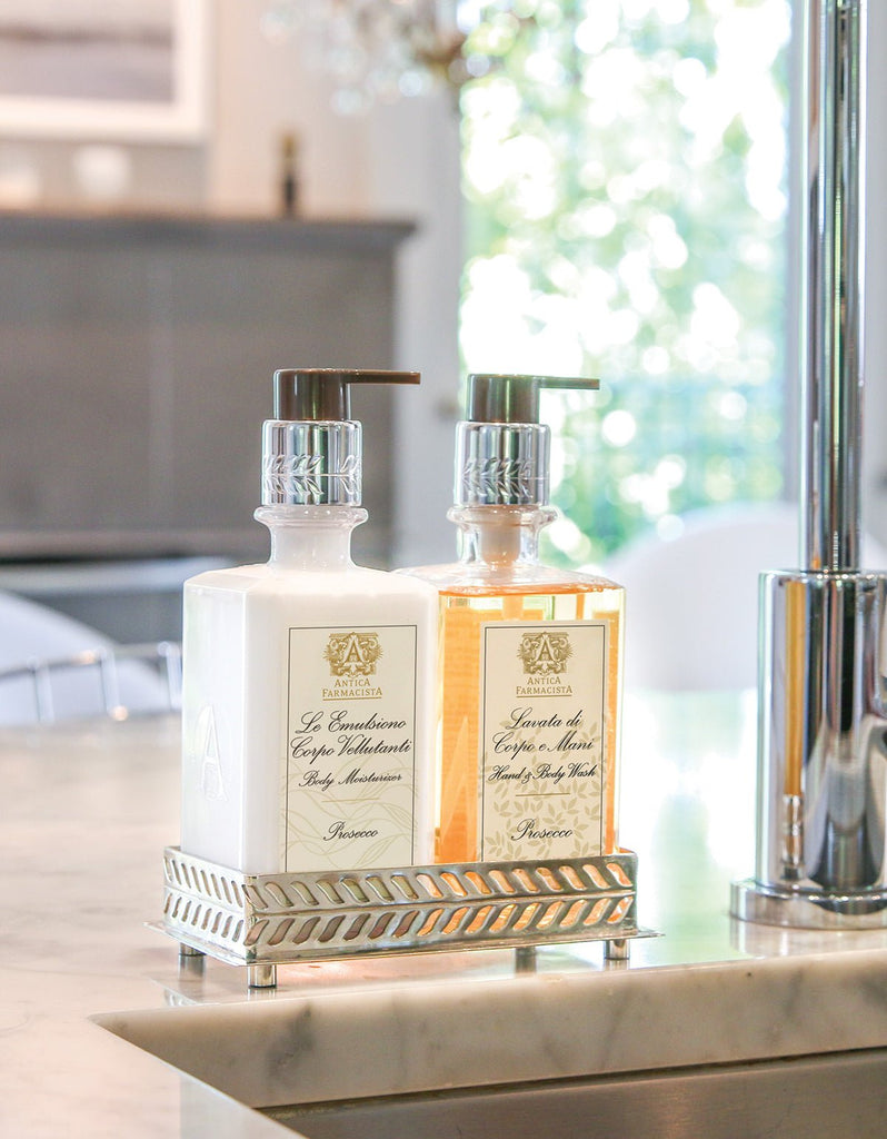 Two decorative soap dispensers rest on a silver tray atop a marble countertop. One contains floral-scented hand & body wash, the other is filled with Antica Farmacista - Prosecco Hand & Body Wash 10oz. The setting appears bright and modern, possibly a kitchen or bathroom.