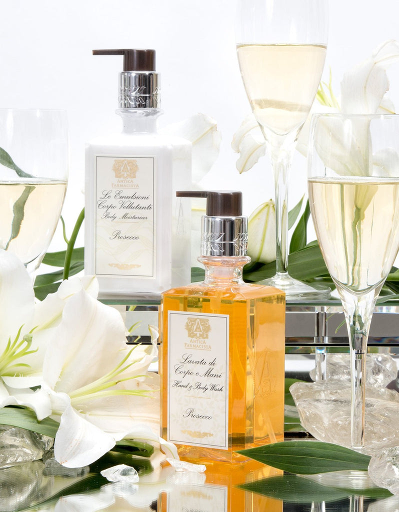 A display of two Antica Farmacista Prosecco Hand & Body Wash bottles with ornate labels, surrounded by white lilies, green leaves, and prosecco glasses. Set on a reflective surface with ice cubes, it evokes the rich fragrance of Madagascar Vanilla.