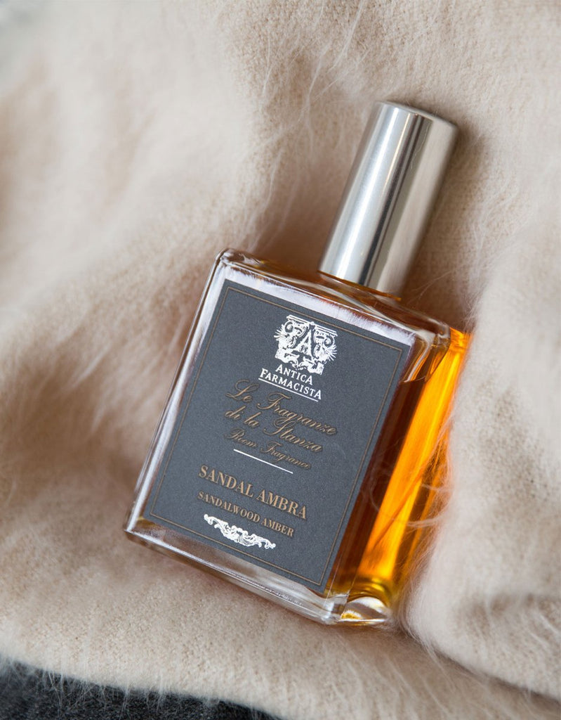 The Antica Farmacista Sandalwood Amber 100ml Room Spray, in a rectangular bottle with a dark label and shiny silver cap, sits on beige fabric. Its sandalwood-amber liquid forms a striking contrast against the creamy background.