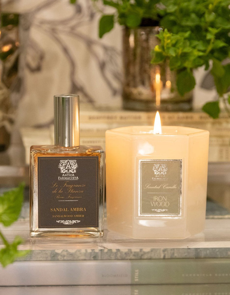 A lit candle labeled Iron Wood sits beside a square bottle of Antica Farmacistas Sandalwood Amber 100ml Room Spray. Both are placed on books with green foliage in the background, creating a cozy and ambient setting reminiscent of an inviting room spray.