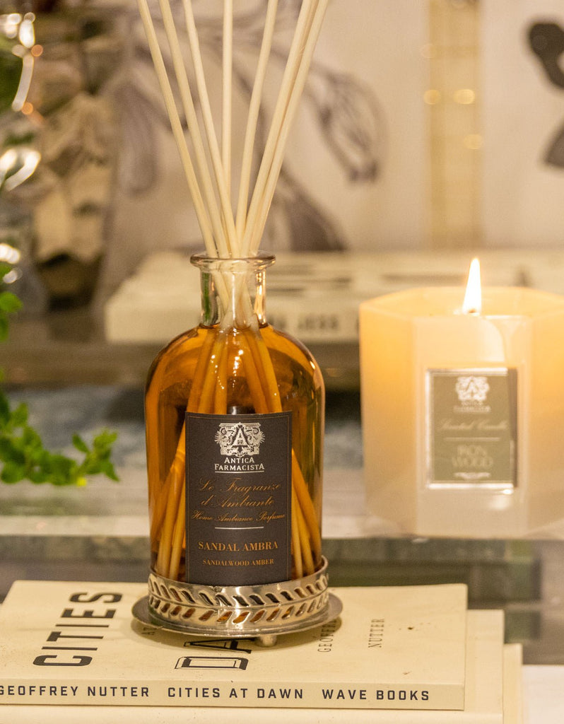 A glass bottle of Antica Farmacistas Sandalwood Amber 250ml Reed Diffuser graces the table. Nearby, a lit beige candle in a glass holder glows warmly. Stacked books Cities at Dawn and Wave Books accompany it, with a soothing plant backdrop amplifying the fragrance.