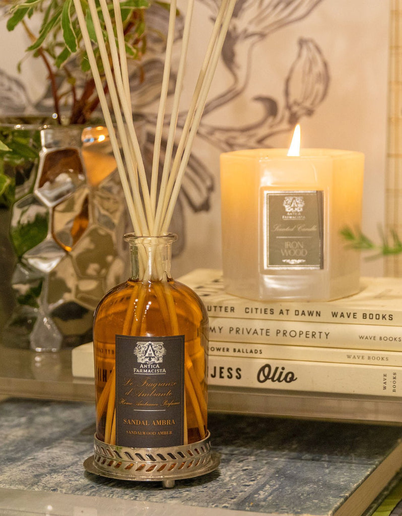 An Antica Farmacista - Sandalwood Amber 250ml Reed Diffuser bottle with Signature Reeds and a lit candle rest on books, while a decorative plant in the background enhances the cozy ambiance.