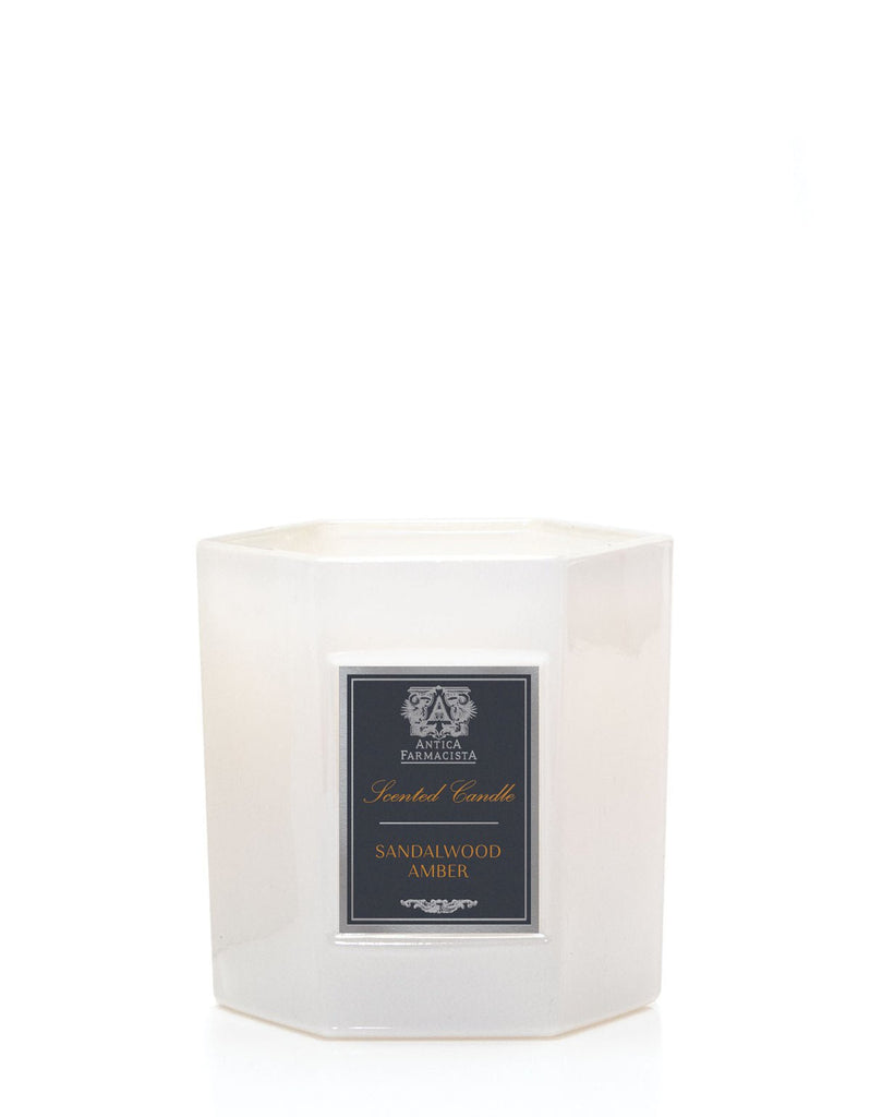 The Antica Farmacista Sandalwood Amber candle features a white hexagonal design with a dark label, reading Scented Candle - Sandalwood Amber, and decorative elements. Made from a soy-paraffin wax blend, it exudes elegance and an inviting aroma against a plain white background.