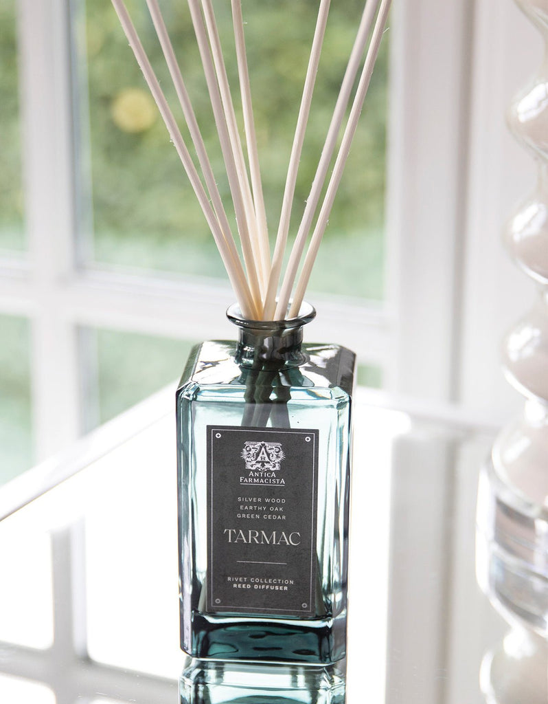 The Antica Farmacista - Tarmac 320ml Reed Diffuser, featuring a dark glass bottle and white reeds, sits on a reflective surface exuding sandalwood fragrance. Sunlight streaming through the window highlights its modern and elegant design.