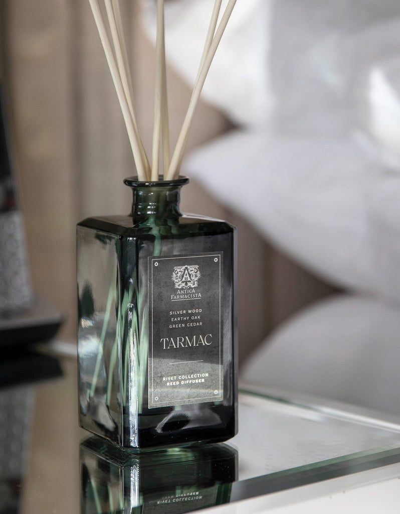 A dark green glass bottle of Antica Farmacistas Tarmac 320ml Reed Diffuser with sticks rests on a reflective surface, featuring notes of Silver Wood, Silky Cedar, and Green Cedar with a hint of sandalwood, part of the exclusive Private Collection.