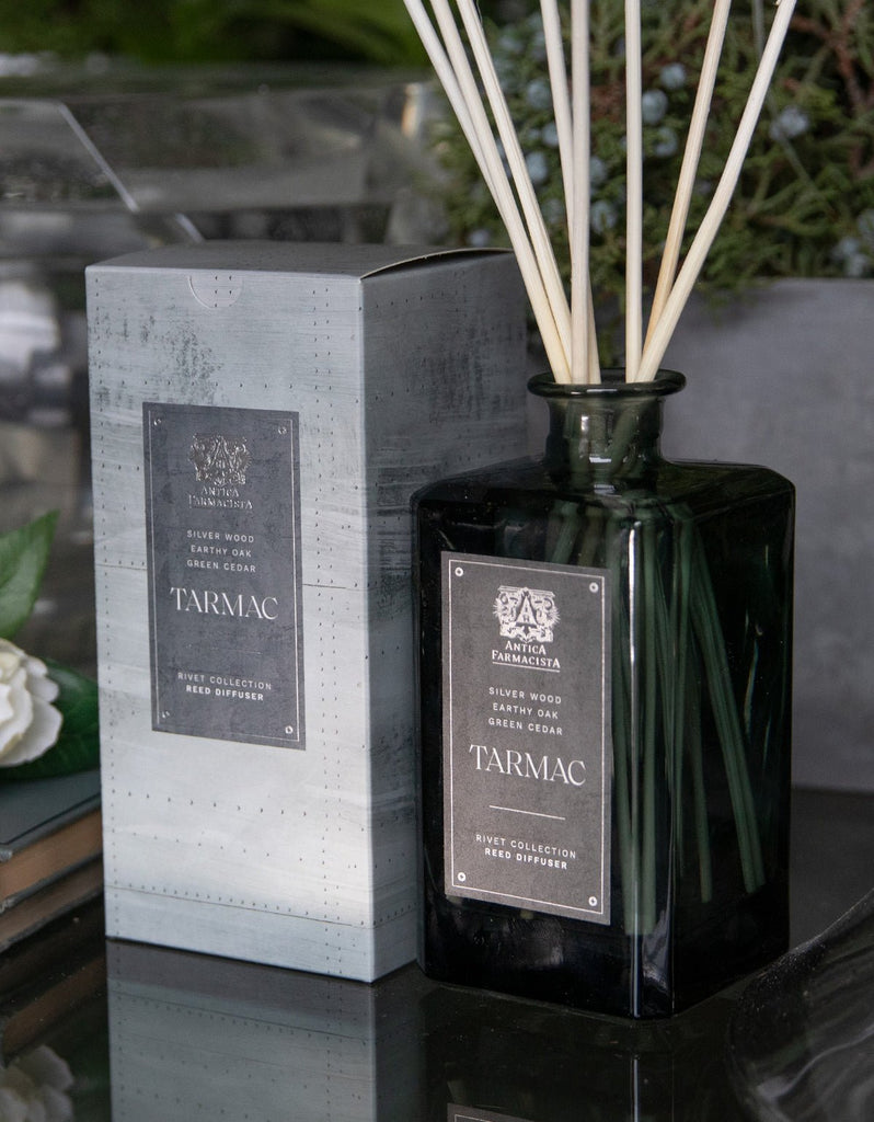 The Antica Farmacista - Tarmac 320ml Reed Diffuser, with a dark green glass bottle and silver Tarmac label, stands by its matching gray box. White reeds diffuse a sandalwood scent amid a greenery backdrop on a reflective surface, enhancing its serene ambiance.