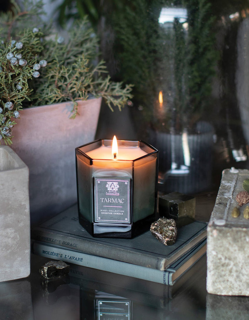 An Antica Farmacista Tarmac 9oz Candle from the Rivet Collection sits on stacked books, surrounded by greenery and stones. Its reflective soy-paraffin wax casts a cozy, serene glow.