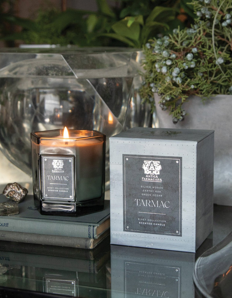 An Antica Farmacista - Tarmac 9oz Candle from the Rivet Collection, crafted with a soy-paraffin blend, sits lit on a book beside its sleek rectangular box. It rests in a glass holder surrounded by greenery and decorative stones, creating an elegant atmosphere.