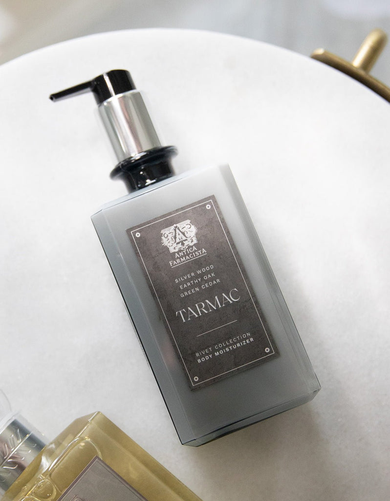 A rectangular pump bottle of Antica Farmacistas Tarmac Body Moisturizer 16oz lies on a white surface. Sporting an ornate emblem, the metallic gray and black label reads Silver Wood, Green Oudh, Green Cedar, combining luxury with natural ingredients for a captivating fragrance.