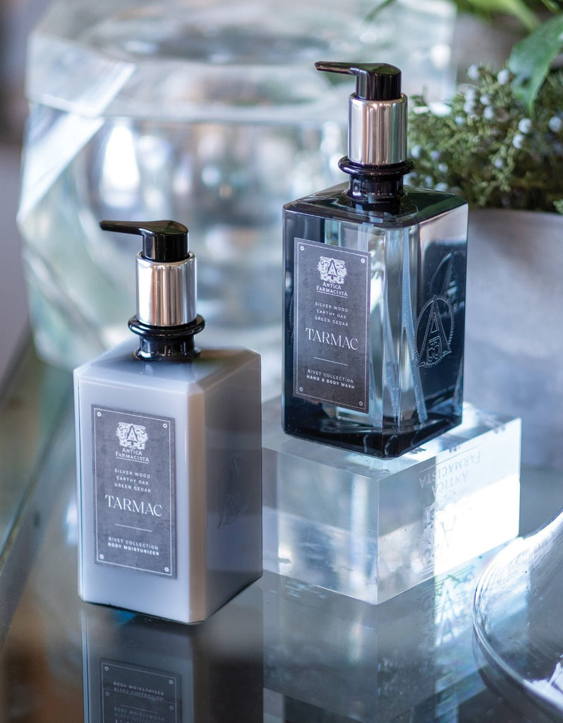 On a reflective surface, two bottles of Antica Farmacistas Tarmac Body Moisturizer with pump dispensers showcase sleek black and silver accents. The moisturizer is enriched with natural ingredients, while greenery in the background adds a refreshing touch.