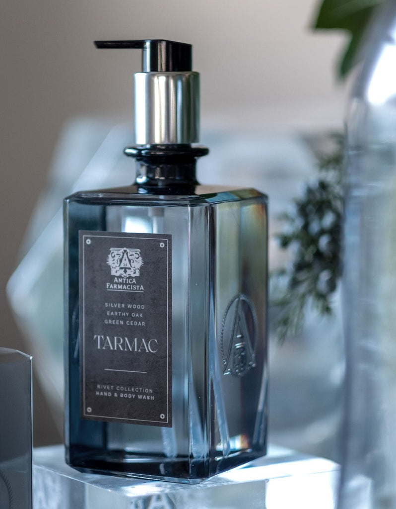 The Antica Farmacista Tarmac Hand & Body Wash 16oz bottle showcases a sleek design with a metallic pump and a label highlighting notes of sitka wood, earthy oak, green cedar, and sandalwood. The background appears softly blurred.