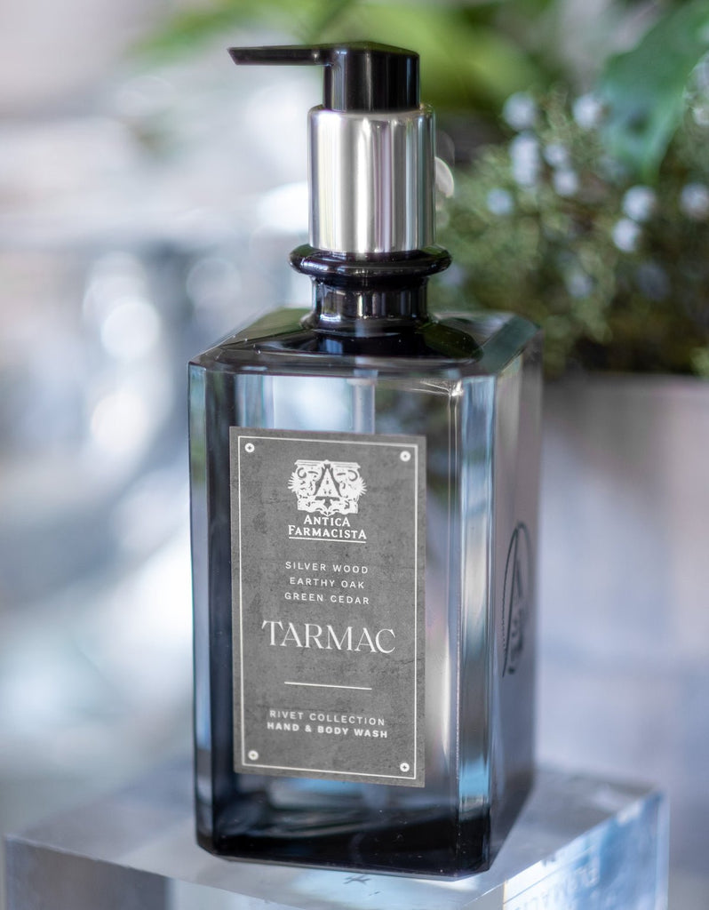 The Antica Farmacista - Tarmac Hand & Body Wash 16oz features a sleek black bottle with a pump dispenser, boasting a blend of sandalwood, earthy oak, and green cedar. Set against a blurred natural backdrop, the bottles elegance is highlighted.