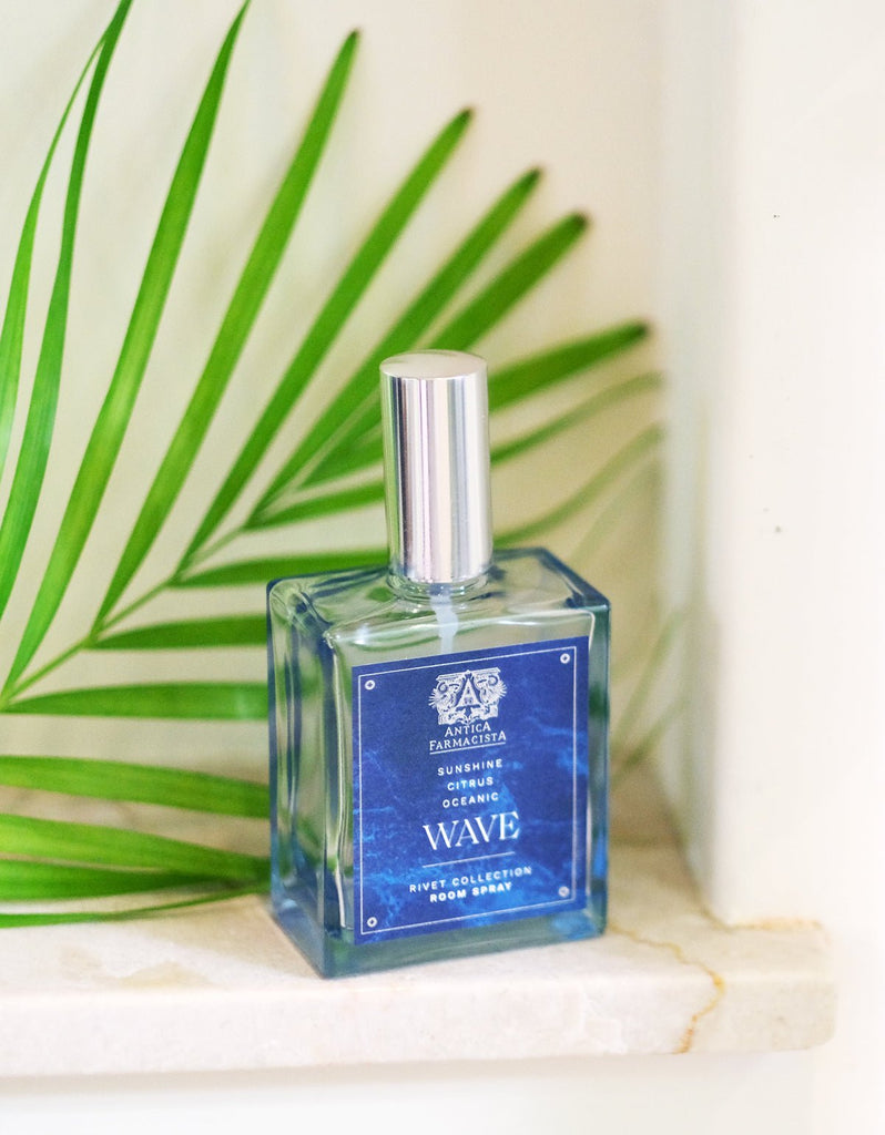 A 100ml glass bottle of Antica Farmacistas Wave Room Spray with a silver cap sits on marble, enhanced by a green fern, adding a fresh touch to the oceanic-themed scene.