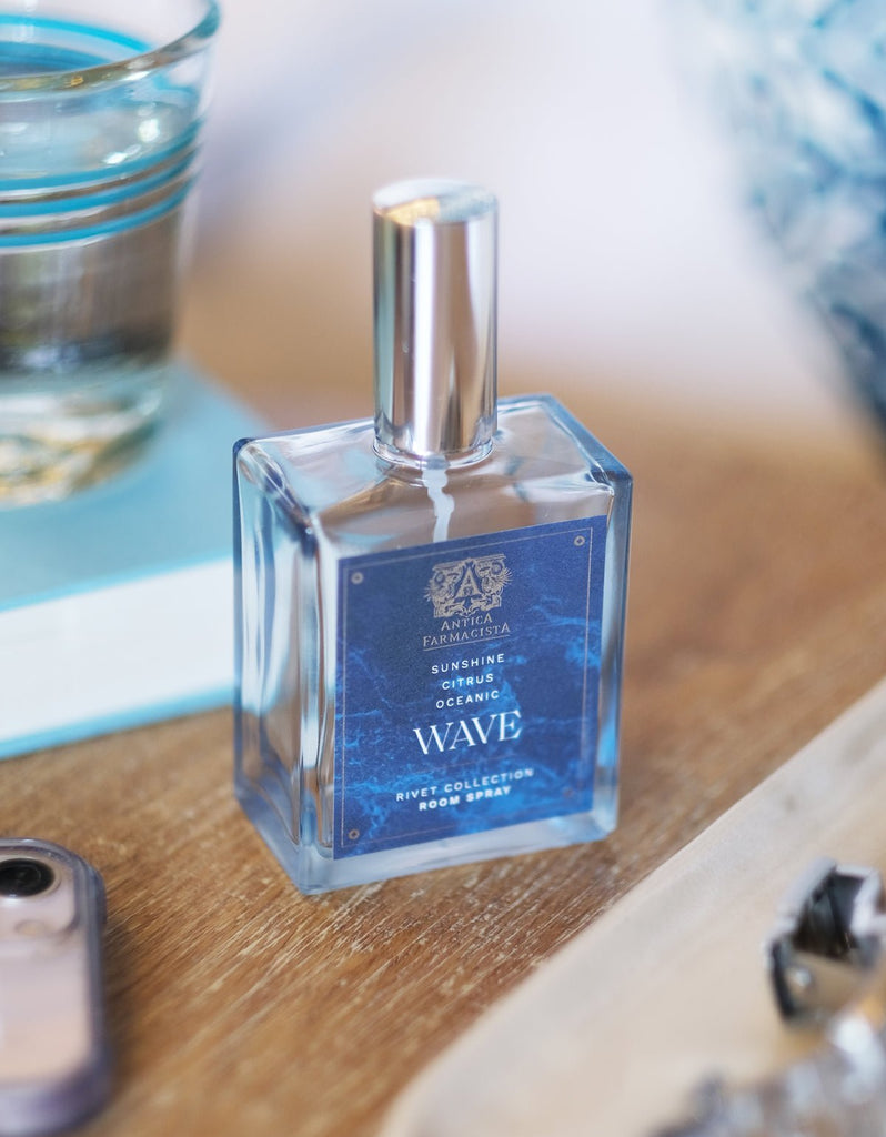 A square glass bottle of Antica Farmacista - Wave 100ml Room Spray from the Rivet Collection sits on a wooden surface, next to a glass of water, a closed book, and a smartphone. Its ocean-themed label embodies refreshing seaside breezes.