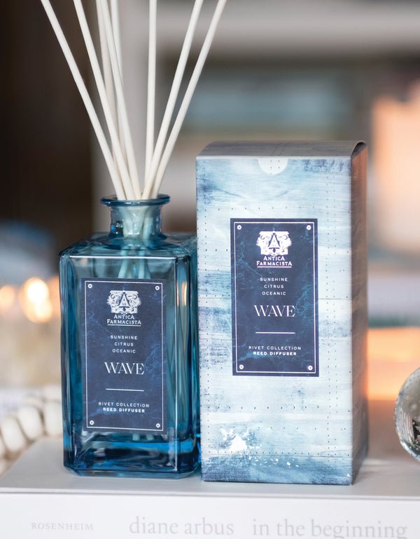 The Antica Farmacista - Wave 320ml Reed Diffuser, in a blue glass bottle, stands elegantly with its matching oceanic design box. Accentuated by marine notes and Meyer Lemon hints, it adds a fresh twist as candles illuminate the blurred decor in the background.