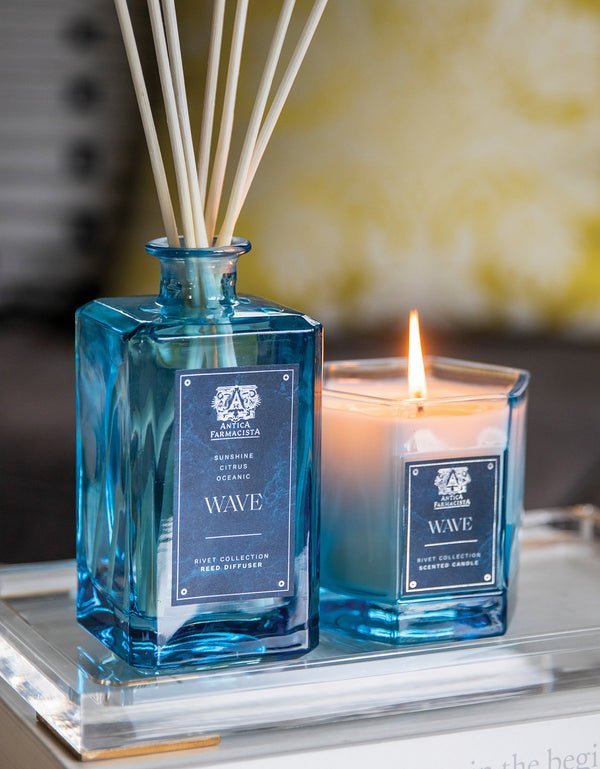 The Antica Farmacista - Wave 320ml Reed Diffuser emits marine notes next to a lit candle from the RYE collection, both labeled Wave. Placed on a tray with a soft-focus backdrop, it evokes the fresh essence of Meyer lemon in each breath.