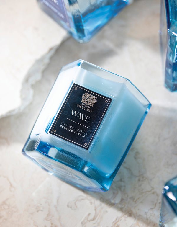 The Antica Farmacista - Wave 9oz blue hexagonal glass candle rests on a light marble surface, releasing a subtle Meyer Lemon scent. Its label bears an intricate emblem above the name. Other similar blue candles can be seen partially in the background.