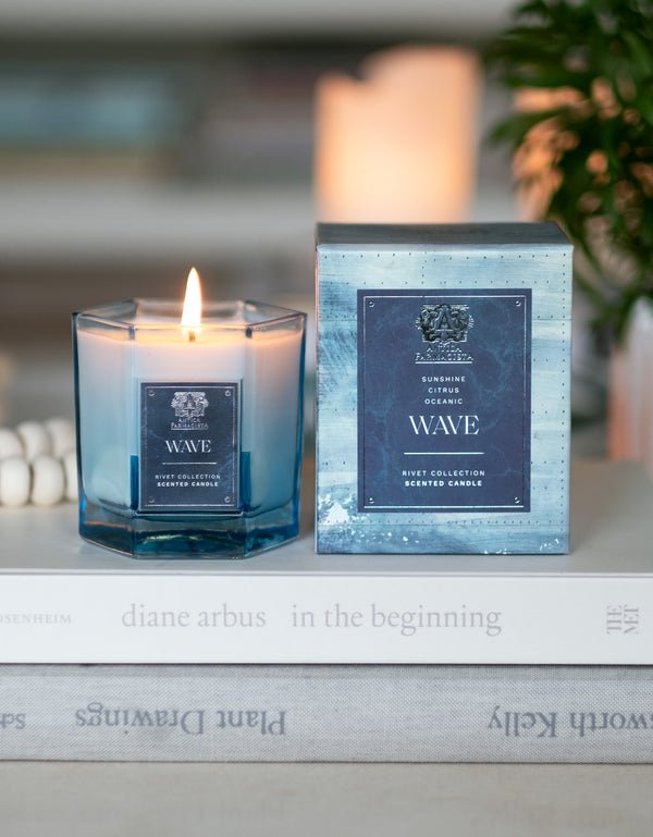 The Antica Farmacista Wave 9oz Candle, crafted from soy-paraffin wax in a blue glass holder, sits beside its matching box on stacked books. The elegant design complements the cozy atmosphere, enhanced by a hint of Meyer lemon fragrance with a blurred plant in the background.