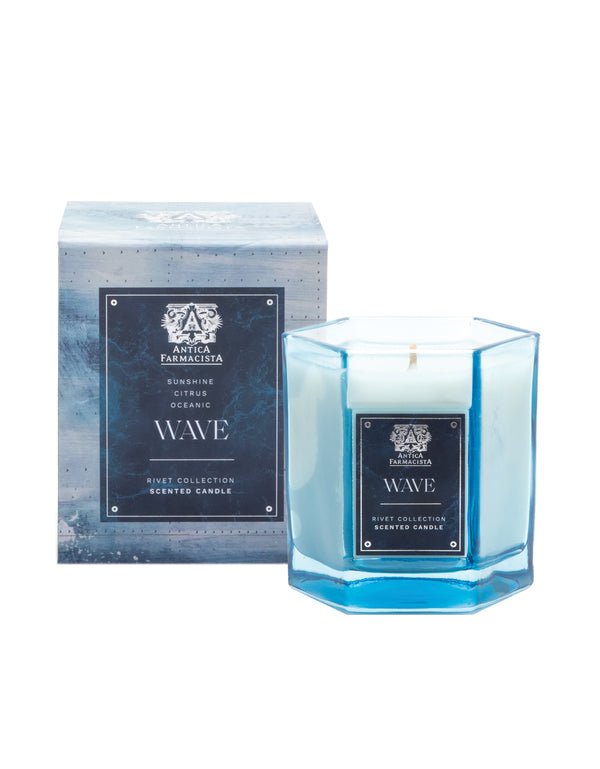 The Antica Farmacista Wave 9oz Candle, featuring an octagonal blue glass design from the Rivet Collection, sits beside its box adorned with oceanic imagery. Described as having a Citrus Oceanic fragrance with Meyer Lemon notes, this Soy-Paraffin Wax blend suits any serene setting.