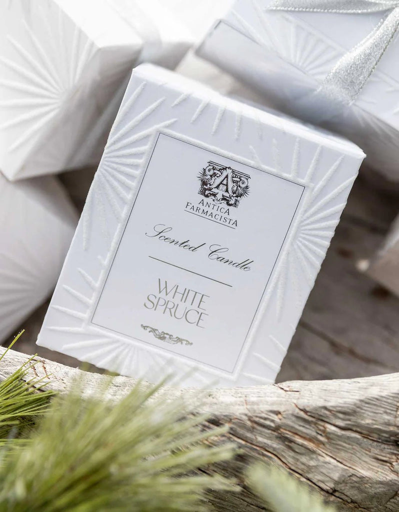 A white box labeled Antica Farmacista - White Spruce 9oz Candle rests on wood, releasing an evergreen scent with green pine needles nearby. The textured design, featuring starburst patterns, hints at the soy-paraffin wax blend inside.