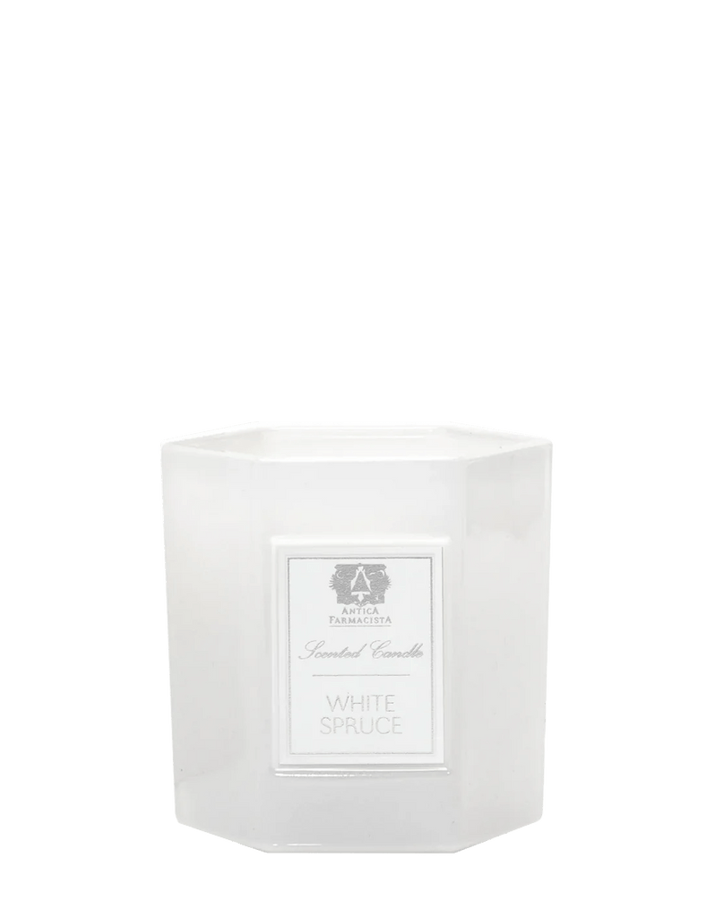 A white scented candle in frosted glass, labeled White Spruce 9oz Candle, stands against a plain backdrop, emitting an evergreen fragrance. Made of a soy-paraffin wax blend, its label also features the brand Antica Farmacista.