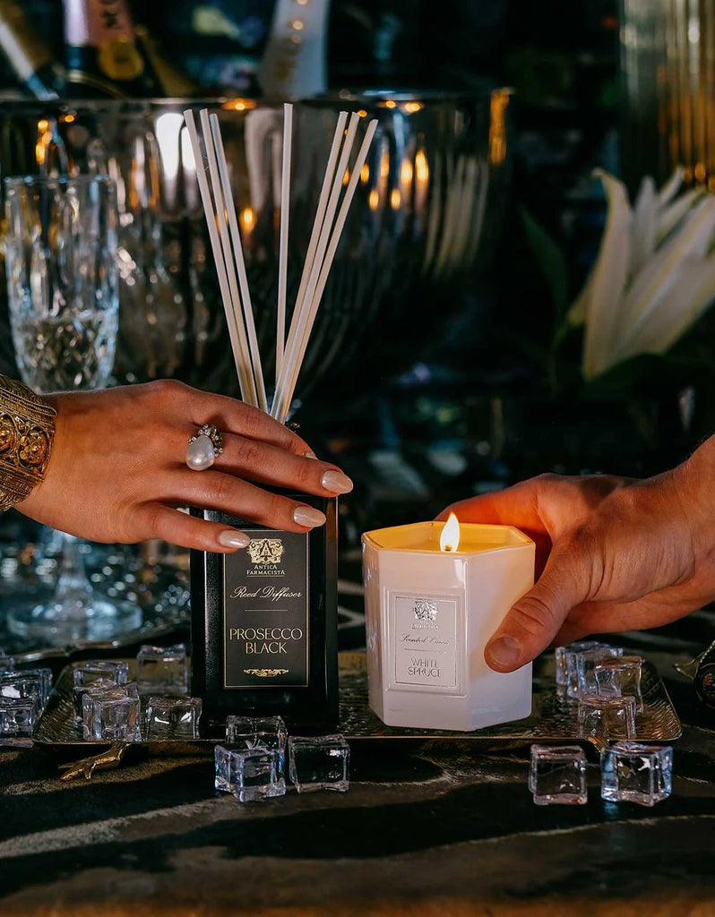 Two hands meet on an elegantly set table. The left holds a Prosecco Black diffuser, the right cradles an Antica Farmacista White Spruce 9oz Candle of soy-paraffin wax. Nearby, ice cubes and glassware enhance the sophisticated ambiance with a hint of evergreen fragrance in the air.