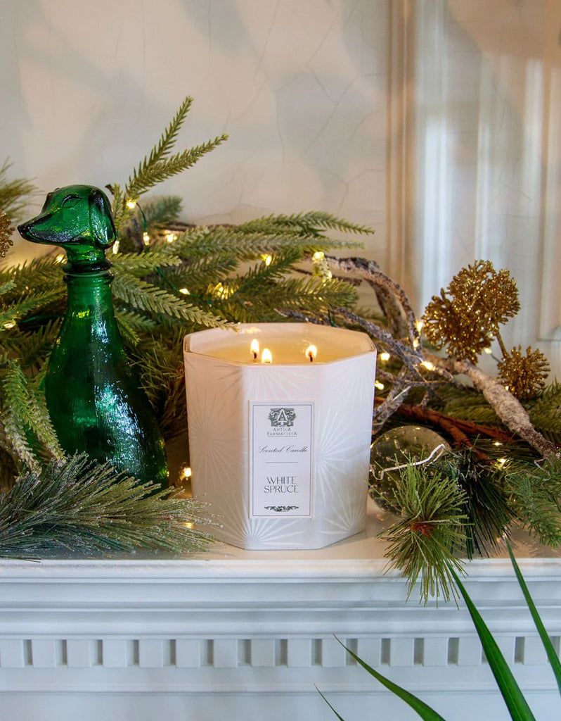 An Antica Farmacista White Spruce Three-Wick Candle graces the mantel, surrounded by fresh spruce branches, a green glass dog-shaped bottle, and golden ornaments, crafting a cozy festive atmosphere.