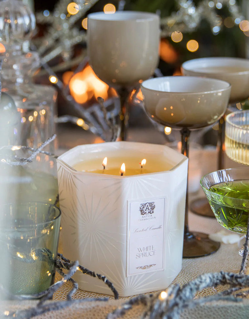 A festive table features the Antica Farmacista - White Spruce Three-Wick Candle, its soft glow illuminating elegant wine glasses and a green dish, all amidst a blurred, warm background.