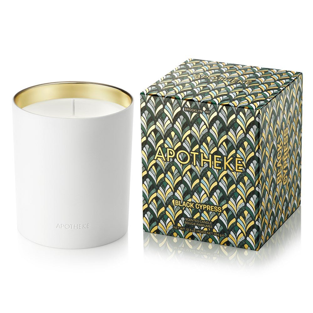 A 12.5oz white ceramic candle by Apotheke, embossed with APOTHEKE, rests beside its decorative box adorned in a green, gold, and blue pattern labeled BLACK CYPRESS, suggesting its aromatic fragrance.