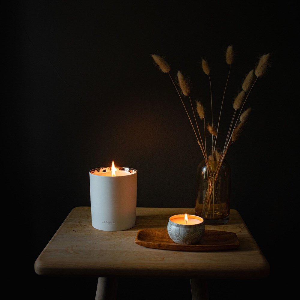 In a dark room, an Apotheke - Black Cypress 12.5oz Candle in a ceramic holder and a small tea light glow on a wooden tray atop a stool. A brown vase with dried grasses sits behind, while the scent of Black Cypress enhances the cozy ambiance bathed in candlelight.