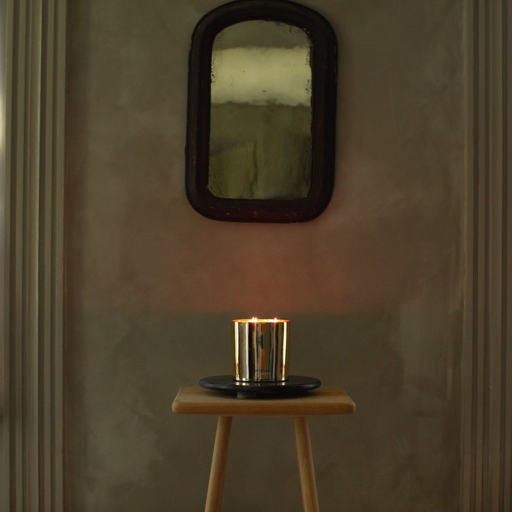 An Apotheke - Black Cypress 3 Wick 72oz Candle in a reflective metallic holder rests on a small wooden stool against a dim wall, emitting an aromatic fragrance. Above it, an antique rectangular mirror with a rounded top is mounted on the wall.