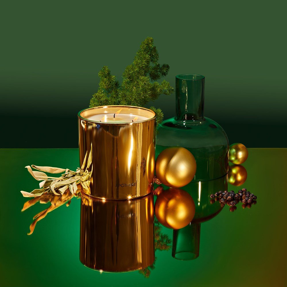 The Apotheke Black Cypress 3 Wick 72oz Candle, featuring a soy wax blend in gold, is surrounded by gold ornaments, a green glass vase, leafy branches, and berries. This elegant setup is displayed against a gradient green background with a reflective surface.