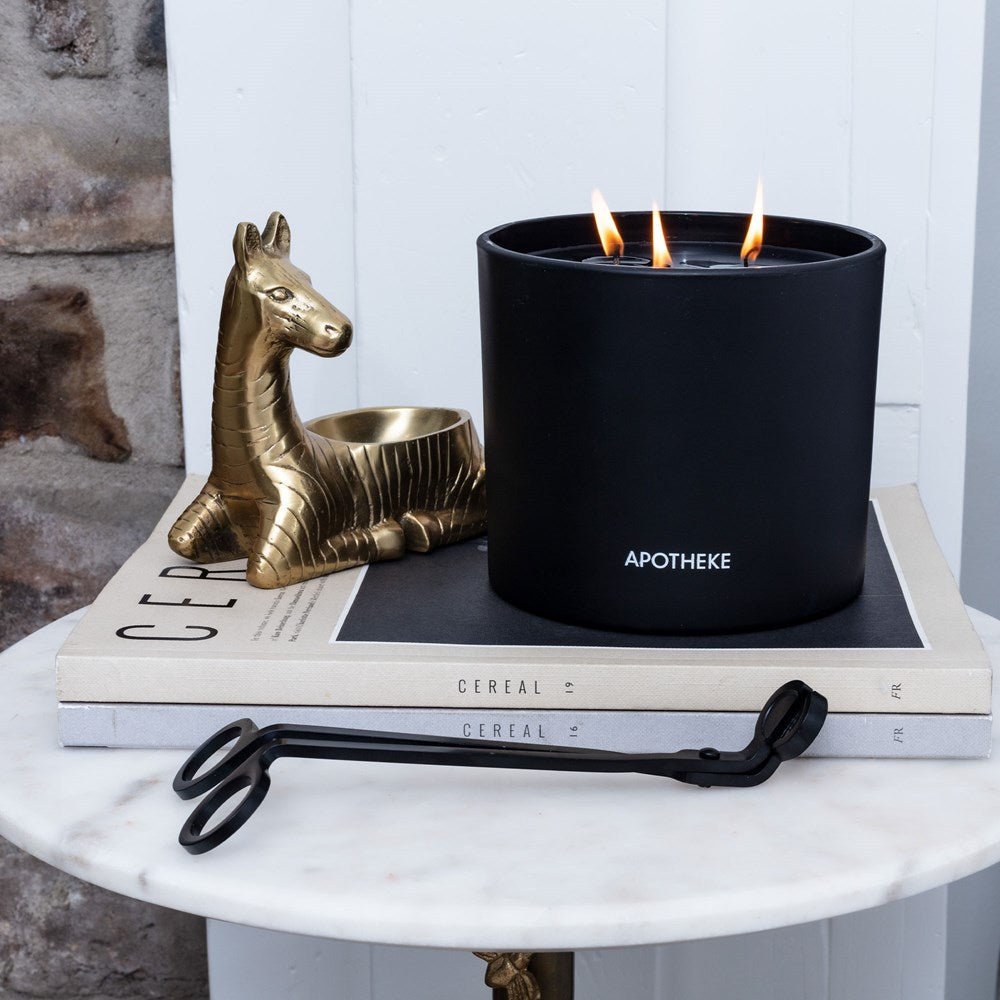 An Apotheke Black candle emits a soothing aroma on a book stack atop a round table. The Apotheke Black Wick Trimmer sits ready for use. Nearby, a gold horse-shaped candle holder enhances elegance against a stone wall and white panel backdrop.