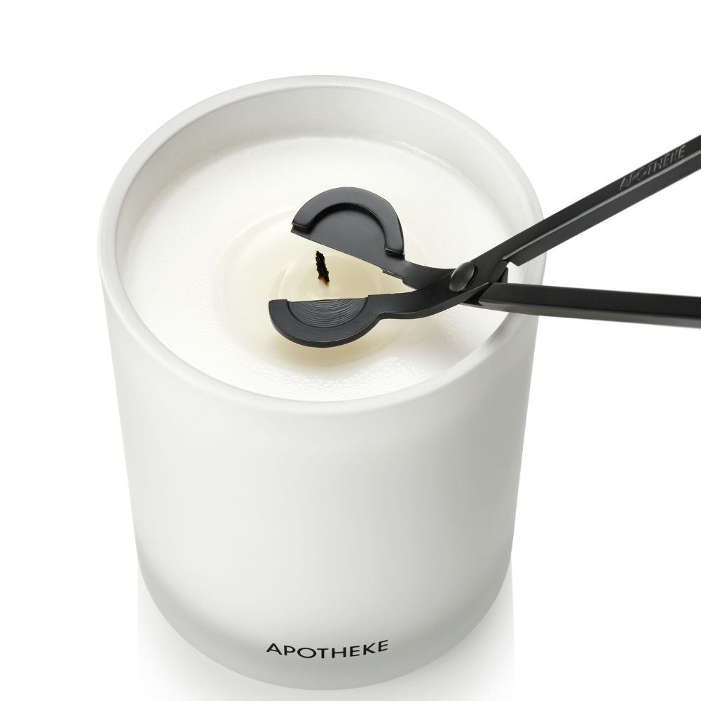 Using the Apotheke Black Wick Trimmer, a blackened wick of a white glass-encased scented candle, labeled APOTHEKE, is carefully trimmed above its smooth white wax for an even burn.