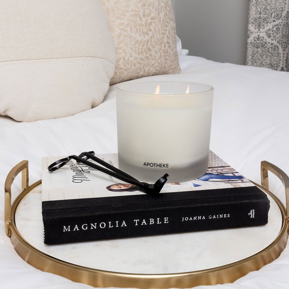 A lit Apotheke candle sits on a closed Magnolia Table book by Joanna Gaines, its aroma wafting gently. A matching Black Wick Trimmer from Apotheke rests beside the glowing candle. All items are neatly arranged on a round marble tray with gold handles atop white linens.