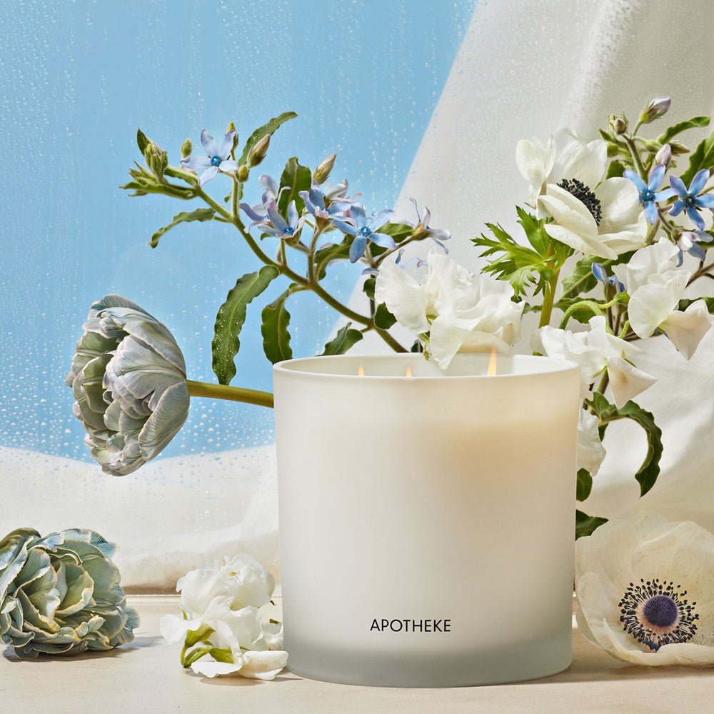 The Apotheke - Canvas 3-Wick Candle in a frosted glass container, labeled APOTHEKE, is surrounded by white and blue flowers. Made from soy wax blend and essential oils, it invites warmth and tranquility against the serene backdrop of a rainy window.