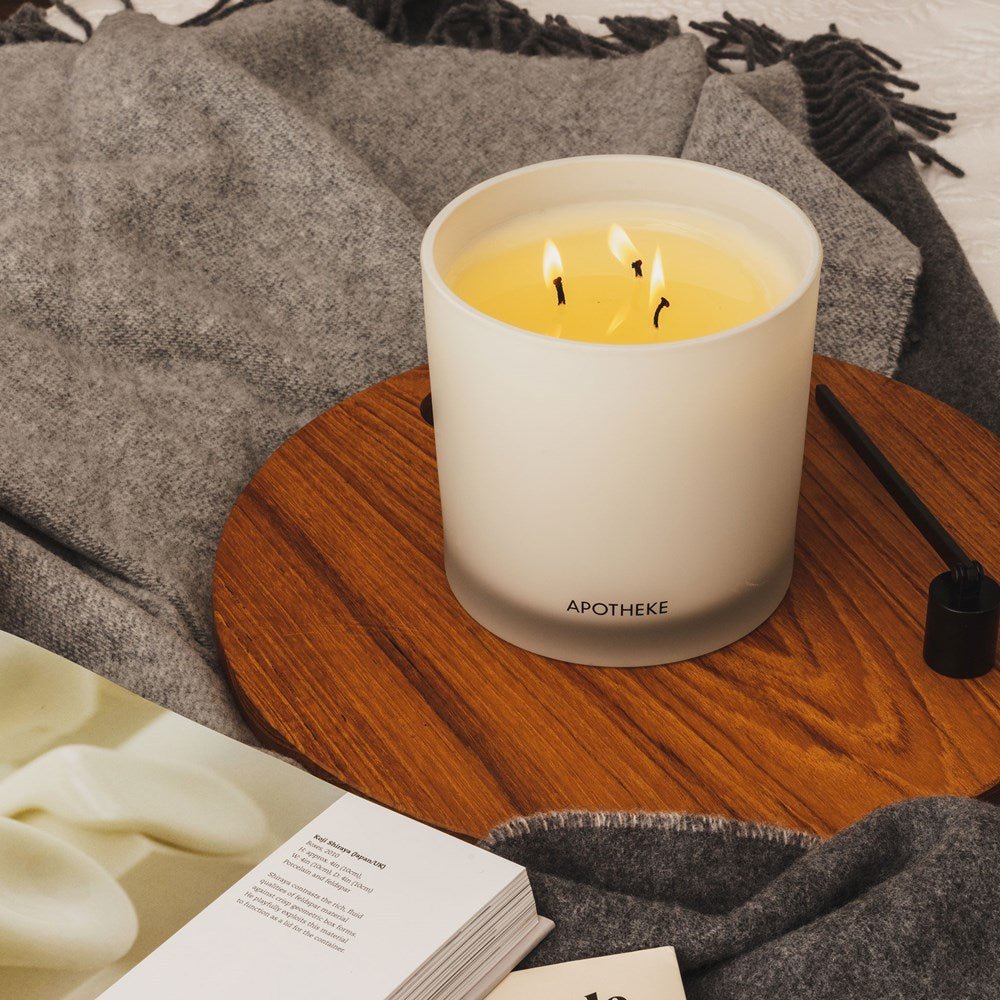 On a wooden tray, an Apotheke Canvas 3-Wick Candle crafted from a soy wax blend flickers beside a soft gray blanket. Nearby, a candle snuffer and an open book add to the cozy setting with essential fragrance oils wafting in the air.