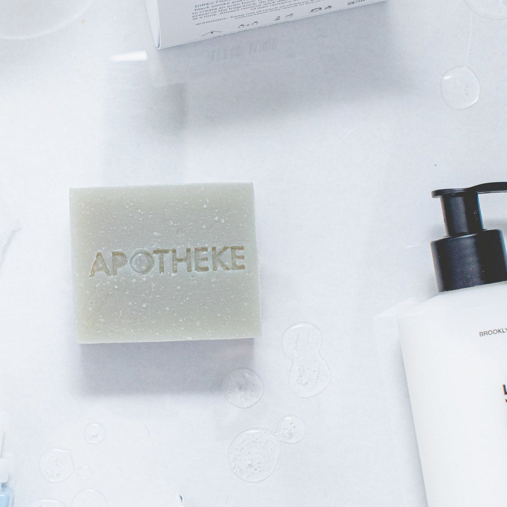 An Apotheke - Canvas Bar Soap with APOTHEKE engraved rests on a white surface, flanked by a partially visible white bottle with a black pump. Small bubbles surround the moisturizing soap.