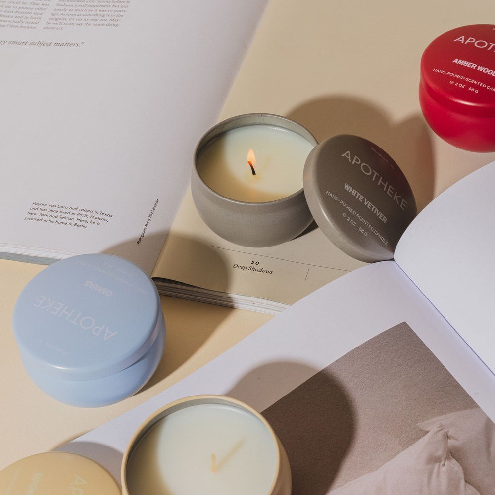 Four Apotheke Canvas Mini Tin 2oz candles in light blue, gray, and red tins sit open next to a book on a cream surface. One is lit, filling the air with delicate floral fragrance from premium soy wax and creating a cozy atmosphere.