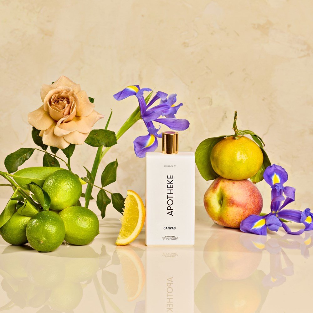 An elegant beige backdrop highlights the Apotheke - Canvas Room Spray alongside a peach rose, purple irises, limes, green apple, lemon slice, and peach. Their glossy reflection enhances the clean and refreshing essential oil fragrance captured in this Apotheke product.