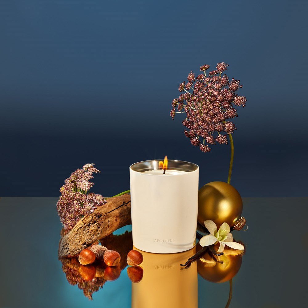 An Apotheke - Cardamom Chestnut 12.5oz Candle sits lit on a reflective surface, surrounded by purple flowers, a gold ornament, hazelnuts, driftwood, and white petals. The air carries its cardamom chestnut gourmand scent against a dark blue gradient backdrop.